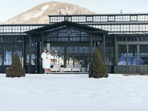 Paar in Bademänteln vor einem Winterhotel mit verschneitem Garten und Bergen