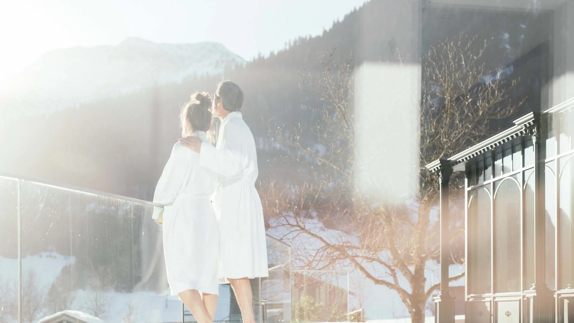 Auf in ein romantisches Hotel in Tirol Paar in Bademänteln genießt sonnige Winterlandschaft auf Balkon