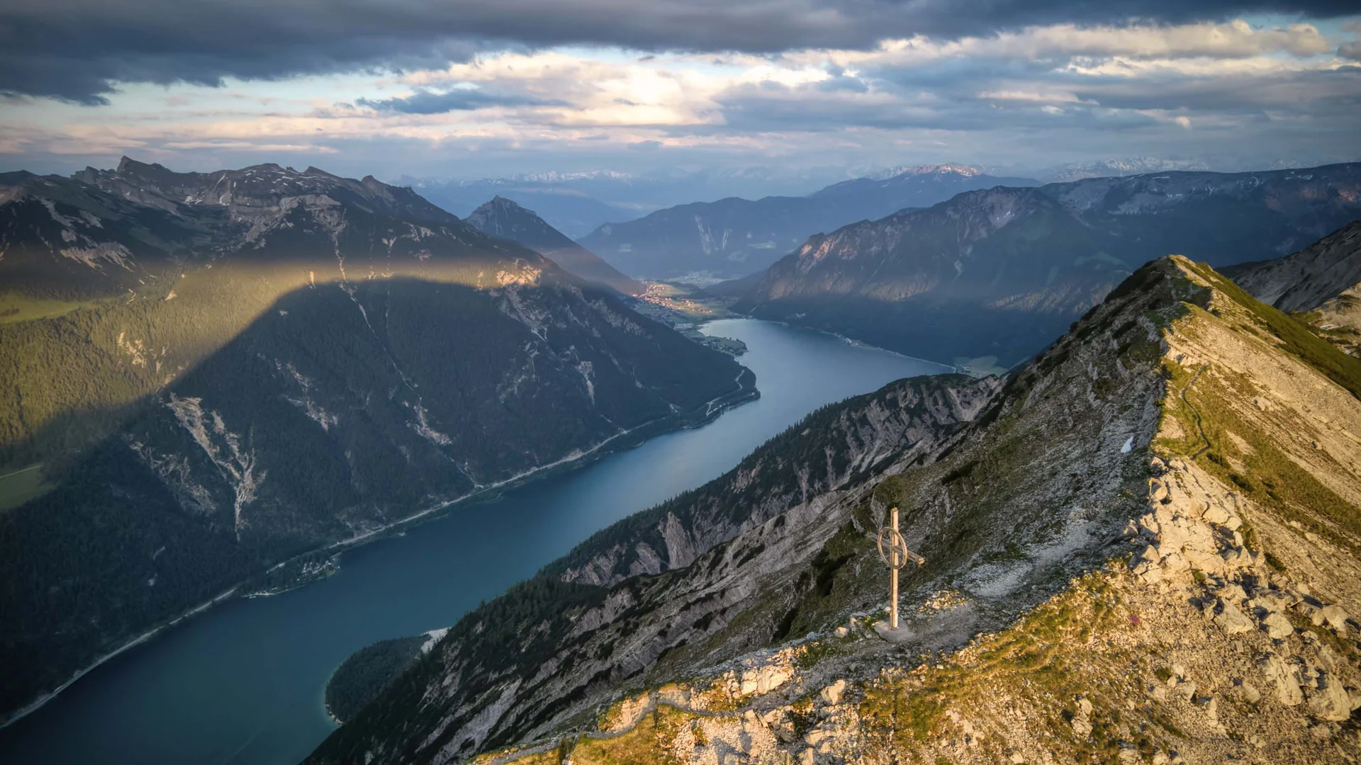 Berggipfel mit Gipfelkreuz und weitem Blick auf See und Berge bei bewölktem Himmel