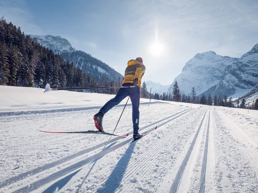 Person beim Langlaufen auf Schneepiste vor verschneiten Bergen bei Sonnenschein