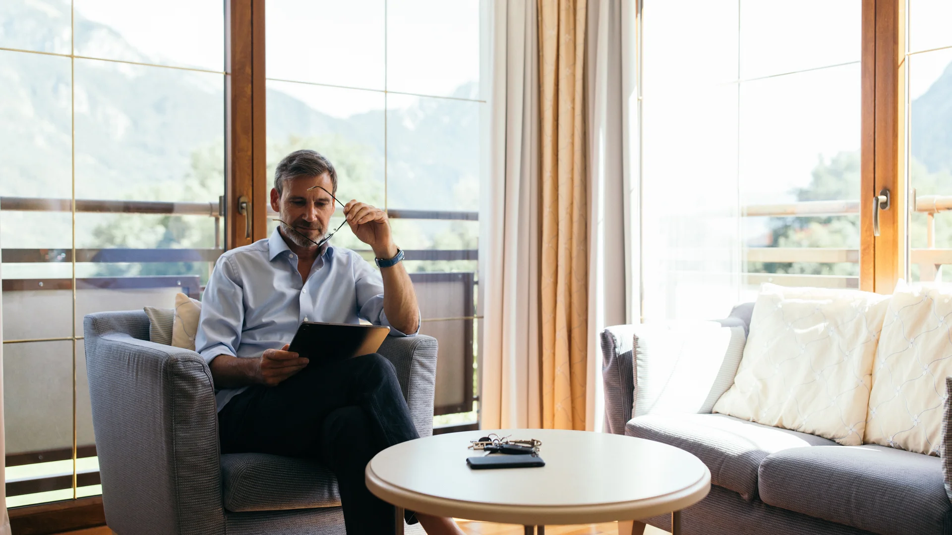 Eure Fragen – unsere Wiesenhof Antworten Mann sitzt auf Sessel und liest Tablet in hellem Wohnzimmer mit Bergblick
