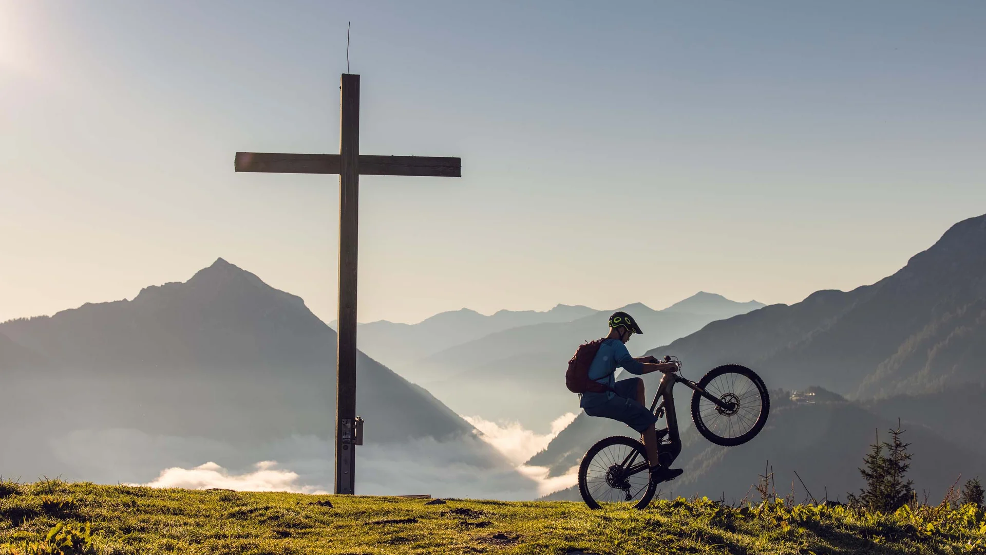 Ganz im Zeichen des Trainings: dieses Sporthotel am Achensee Mountainbiker macht Wheelie neben Gipfelkreuz mit Bergpanorama im Hintergrund