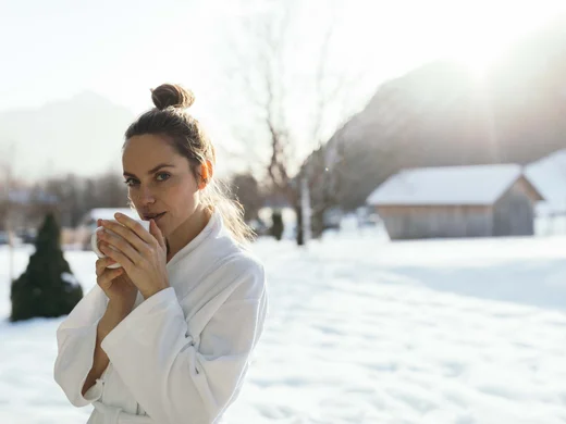 Auszeit im Single-Hotel in Österreich Frau in Bademantel trinkt heißes Getränk draußen im Schnee vor Hütte