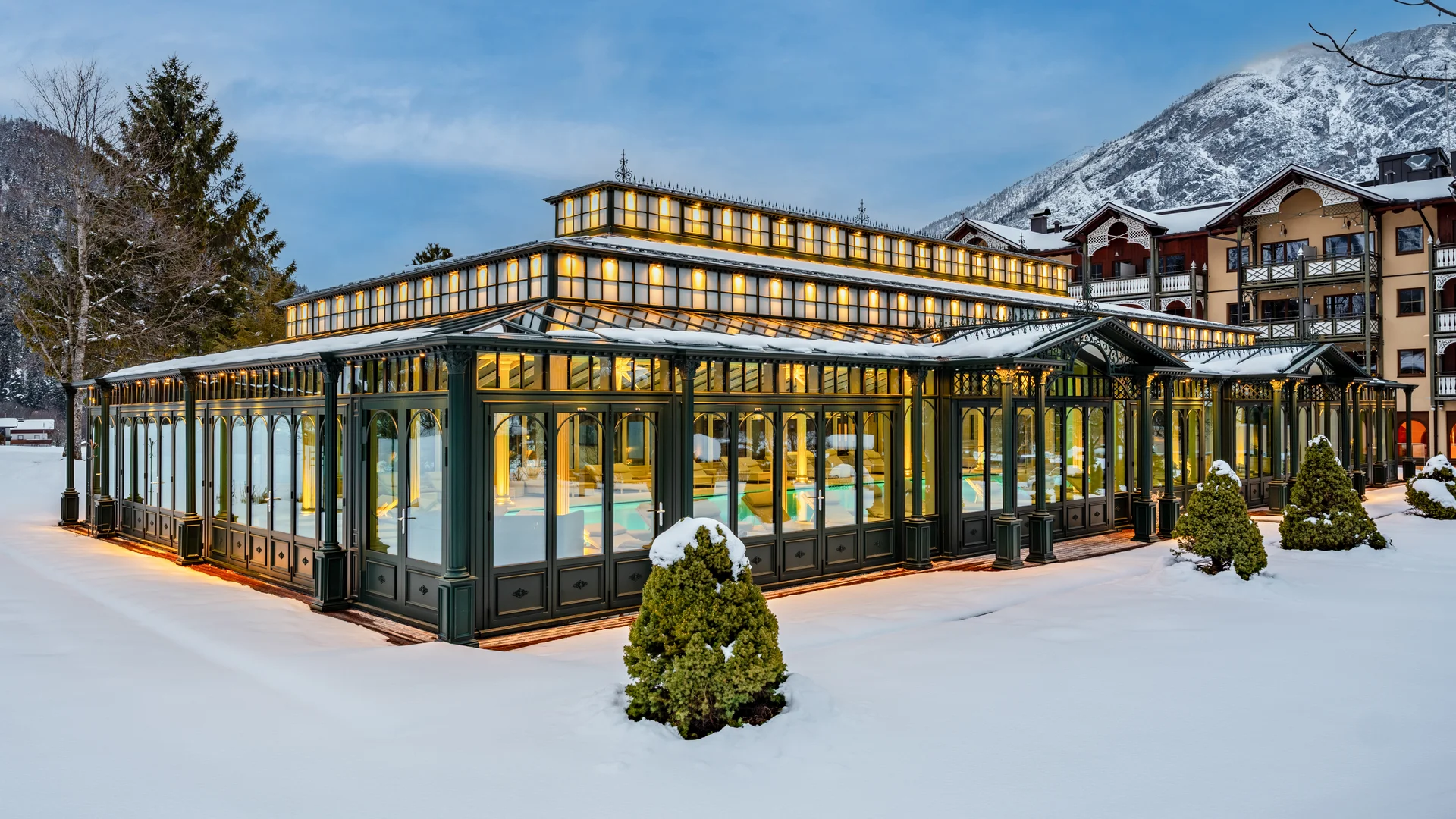 Beleuchteter gläserner Pavillon in verschneiter Winterlandschaft vor Bergen