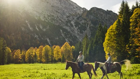 Two riders on horses in a green mountain meadow with colorful autumn trees