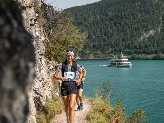 Frau läuft am Seeufer entlang bei einem Rennen mit Boot und bewaldetem Hügel im Hintergrund