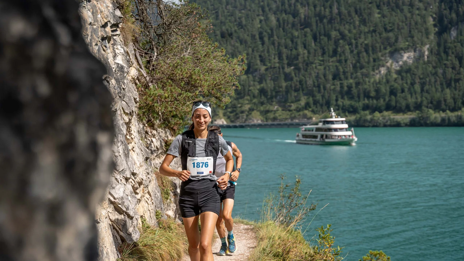Frau läuft am Seeufer entlang bei einem Rennen mit Boot und bewaldetem Hügel im Hintergrund