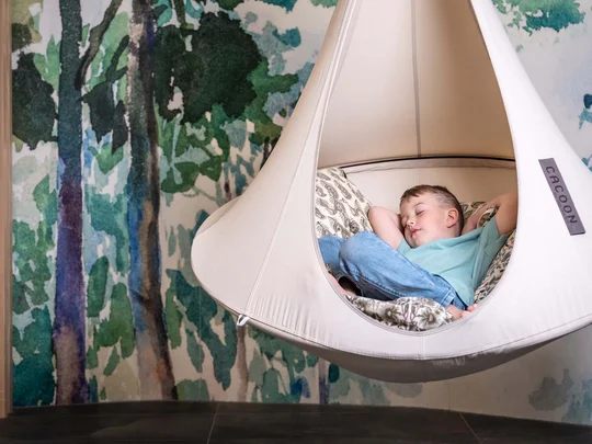 Eure Fragen – unsere Wiesenhof Antworten Kind schläft in einer hängenden Kokon-Wiege vor einer Waldtapete