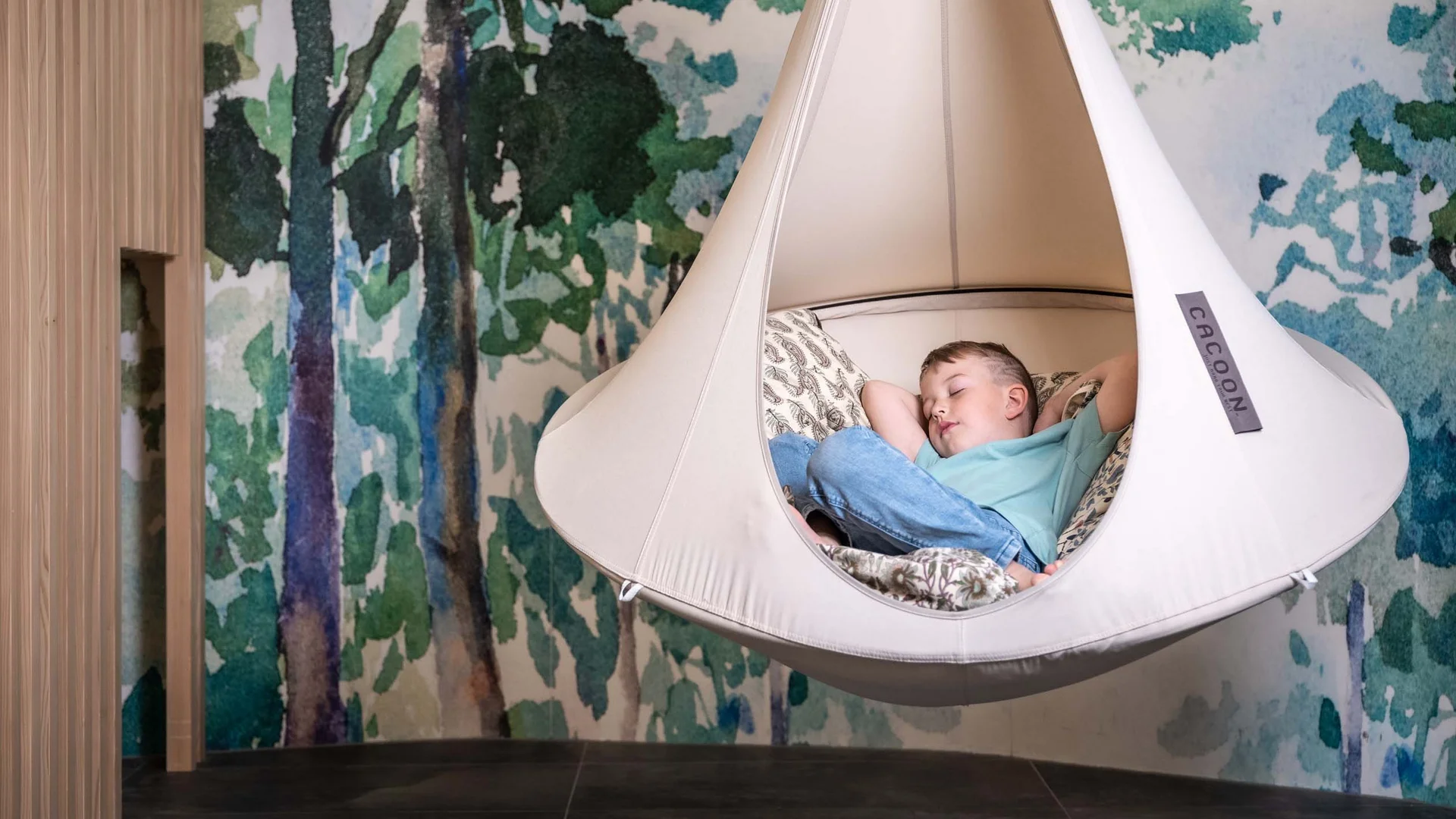Euer Familienhotel am Achensee Kind schläft in einer hängenden Kokon-Wiege vor einer Waldtapete