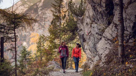 Zwei Frauen wandern auf einem Bergweg mit Felsen und Bäumen im Herbst