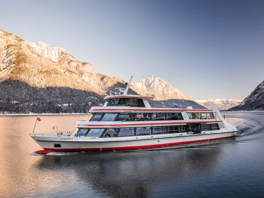 In, auf und rund um den Achensee Passagierschiff auf ruhigem Bergsee mit schneebedeckten Bergen bei Sonnenuntergang
