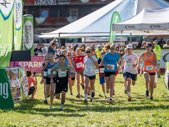 Kinder laufen bei einem Rennen auf einer sonnigen Wiese mit Zuschauern im Hintergrund