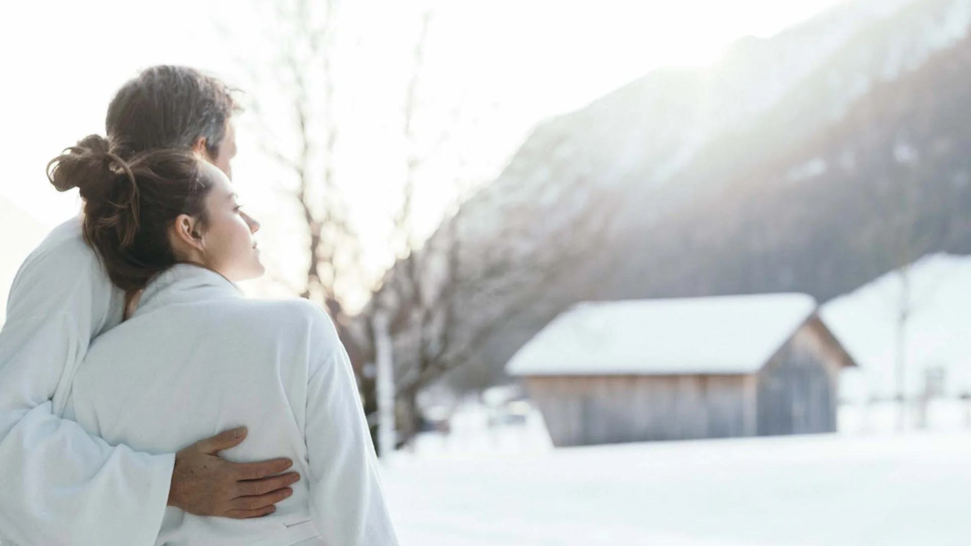 Auf in ein romantisches Hotel in Tirol Paar in Bademänteln umarmt sich vor verschneiter Landschaft mit Hütten
