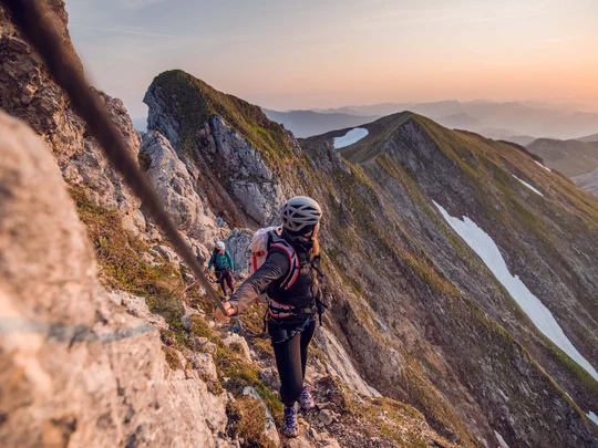 Wanderhotel Tirol: durchs Karwendel wandern und biken Kletterer sichern sich an Seil an steiler Felswand bei Sonnenuntergang in den Bergen