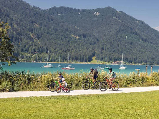 Euer Familienhotel am Achensee Familie fährt mit Fahrrädern am See entlang vor bewaldeten Bergen und Segelbooten