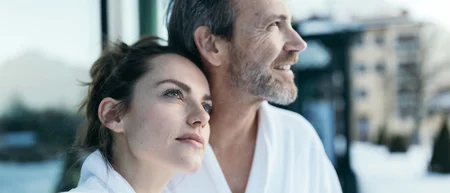 Paar in weißen Bademänteln schaut entspannt in die Ferne im Schnee