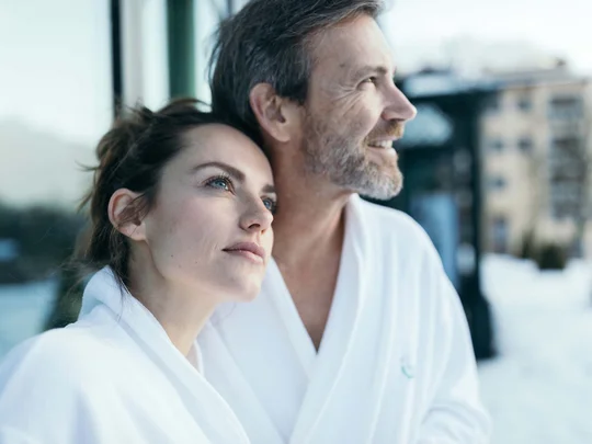 Auf in ein romantisches Hotel in Tirol Paar in weißen Bademänteln schaut entspannt in die Ferne im Schnee
