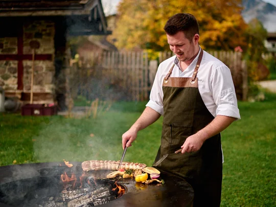 Das 4-Sterne-Hotel am Achensee: der Wiesenhof Mann mit Schürze grillt Fleisch und Gemüse draußen auf Feuerstelle