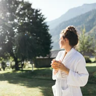 Frau im weißen Bademantel trinkt Tee im Garten mit Bergblick am Morgen