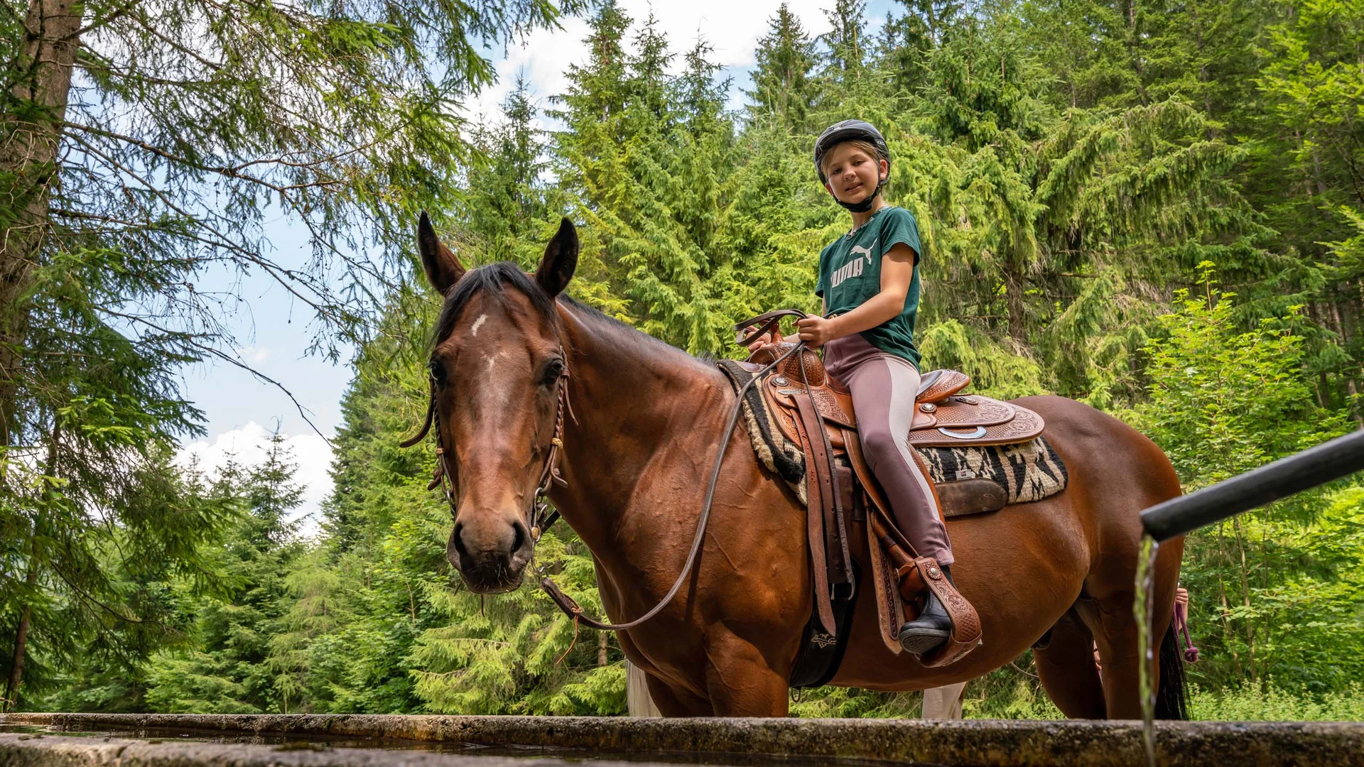 Familienhotel mit Ponyreiten am Achensee | Wiesenhof Pertisau Kind mit Helm reitet braunes Pferd im Wald neben einem Wasserbecken