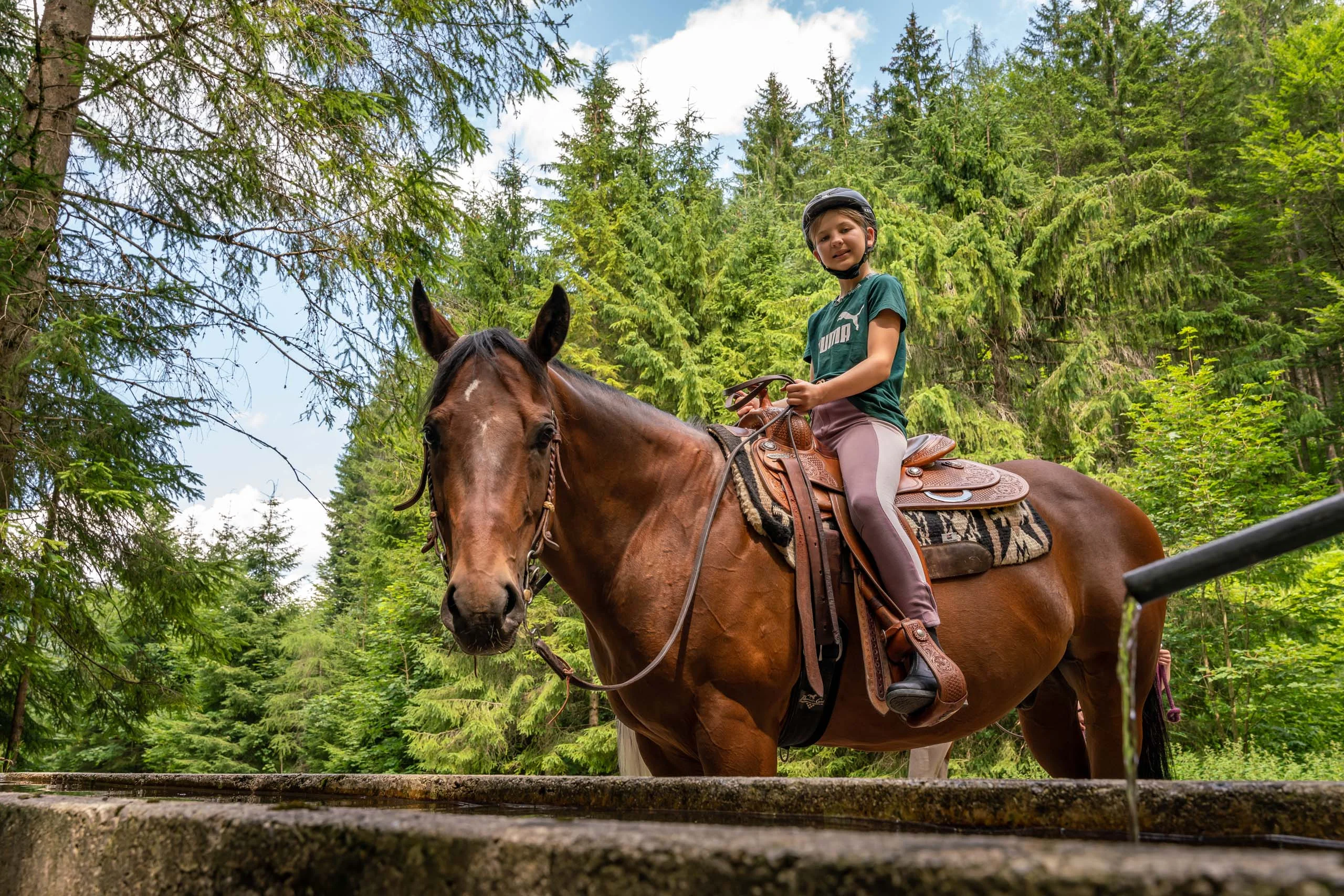 Familienhotel mit Ponyreiten am Achensee | Wiesenhof Pertisau Kind mit Helm reitet braunes Pferd im Wald neben einem Wasserbecken