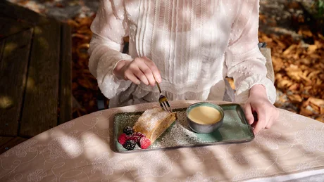 Person in weißer Bluse isst Kuchen mit Beeren und trinkt Kaffee im Freien