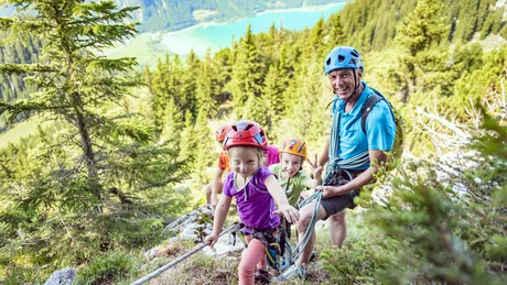 Kinder und Erwachsener klettern mit Helmen und Seilen an einem bewaldeten Berg
