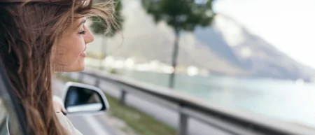 Junge Frau mit wehendem Haar blickt aus Autofenster auf See und Berge