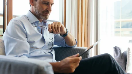 Mann sitzt auf Sofa und liest Tablet, hält Brille in der Hand