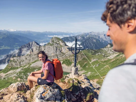 Sehenswürdigkeiten am Achensee rund um den Wiesenhof Frau mit rotem Rucksack sitzt auf Berggipfel vor Gipfelkreuz, Mann schaut zu ihr