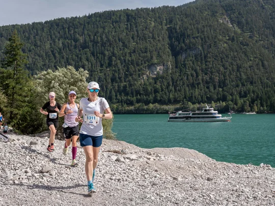 Läuferinnen laufen auf einem felsigen Weg am See mit Boot und bewaldeten Bergen im Hintergrund