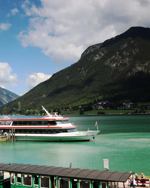 Grüner historischer Zug und Ausflugsboot am See mit Bergen und blauem Himmel