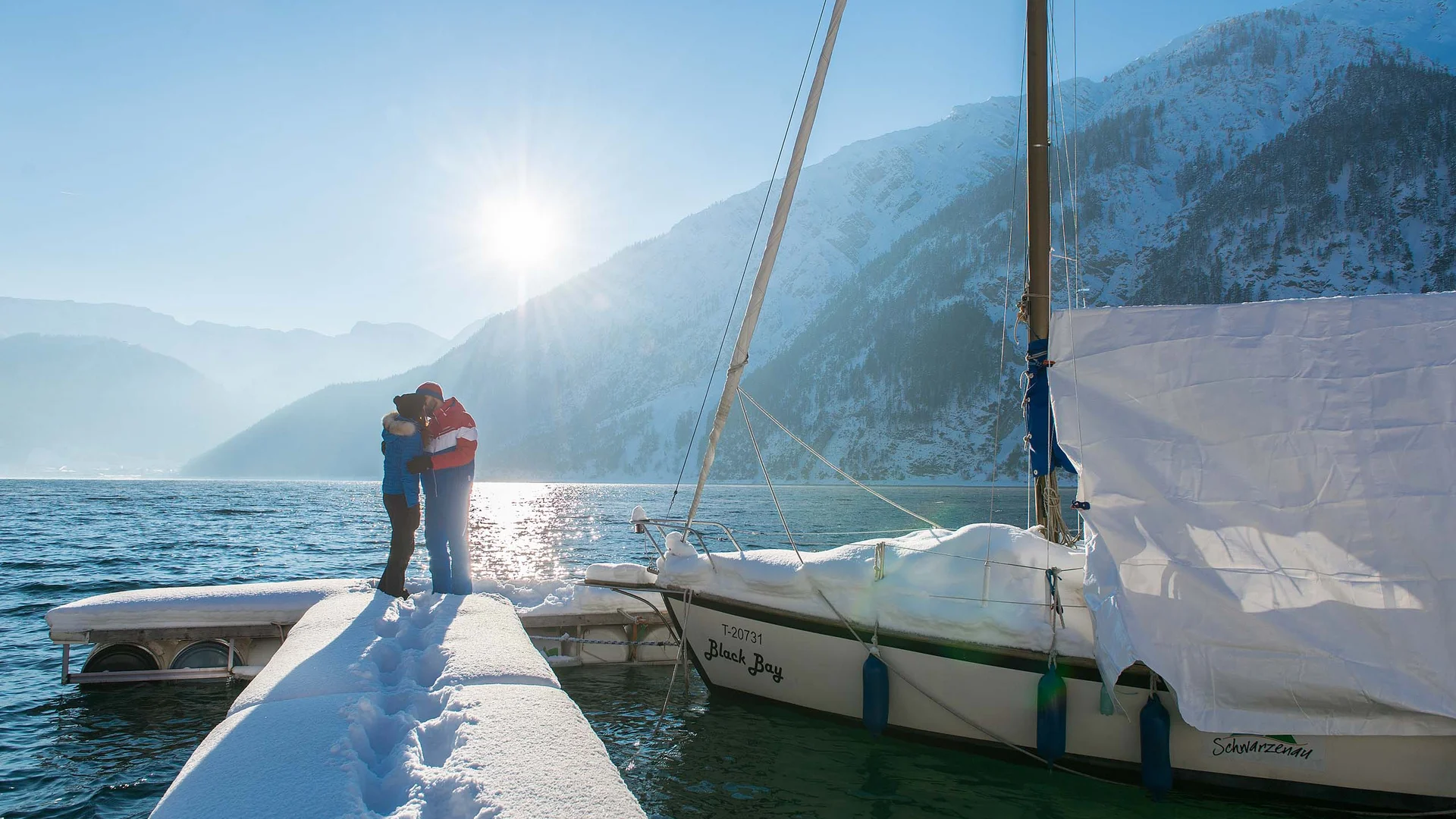 Wanderhotel Tirol: durchs Karwendel wandern und biken Paar umarmt sich auf schneebedecktem Steg neben Segelboot im winterlichen Bergsee
