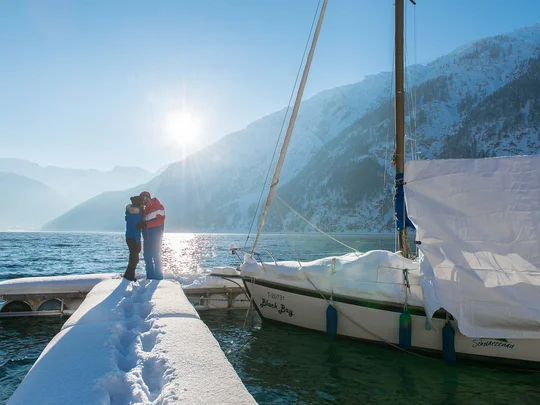 Wanderhotel Tirol: durchs Karwendel wandern und biken Paar umarmt sich auf schneebedecktem Steg neben Segelboot im winterlichen Bergsee
