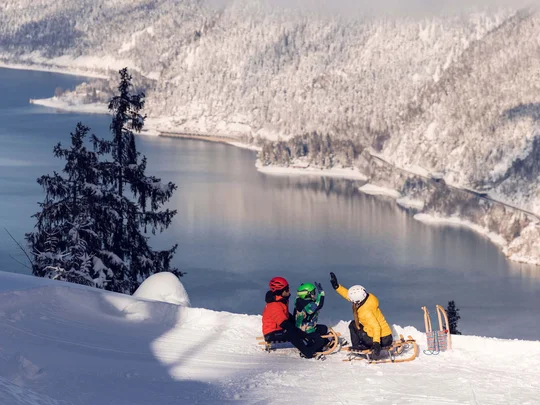 Euer Familienhotel am Achensee Drei Personen beim Rodeln auf schneebedecktem Hang mit Bergsee im Hintergrund