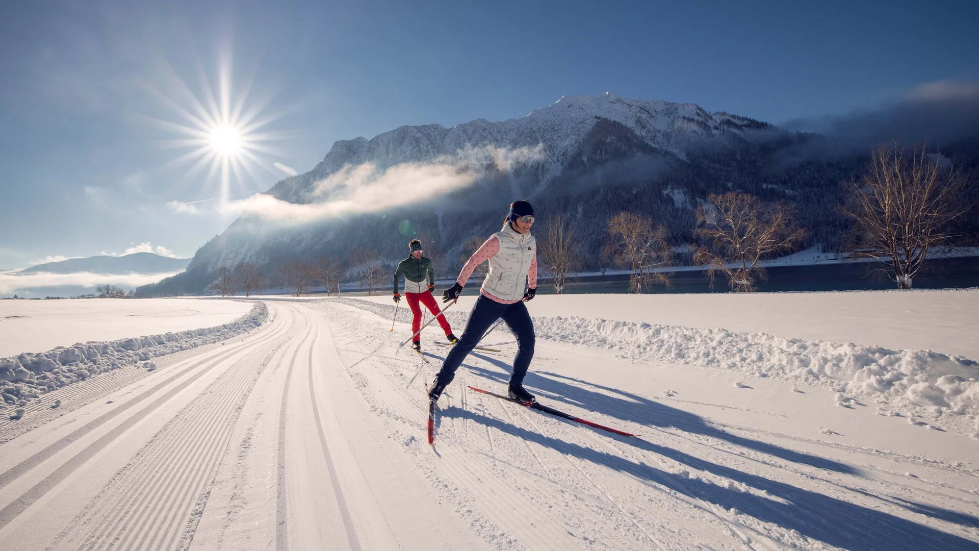 Zwei Personen beim Langlaufen auf verschneiter Strecke vor Bergkulisse bei Sonne