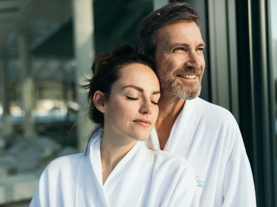 Paar in weißen Bademänteln entspannt draußen vor Glaswand