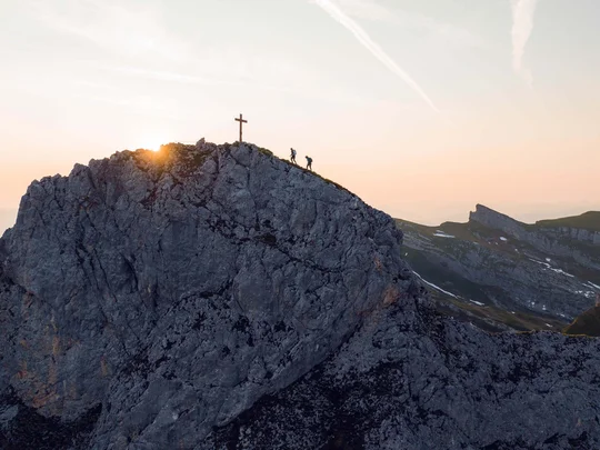 Wanderhotel Tirol: durchs Karwendel wandern und biken Wanderer steigen auf einen felsigen Berggipfel mit Kreuz bei Sonnenuntergang