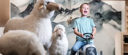 Lachender Junge auf Spielauto neben drei hellen Schaffiguren im Raum