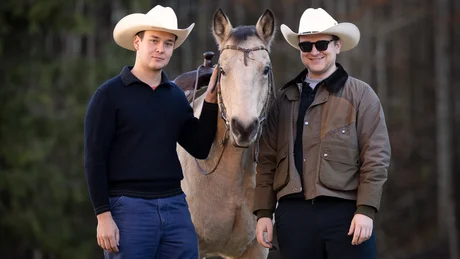 Two men in cowboy hats standing next to a saddled horse outdoors