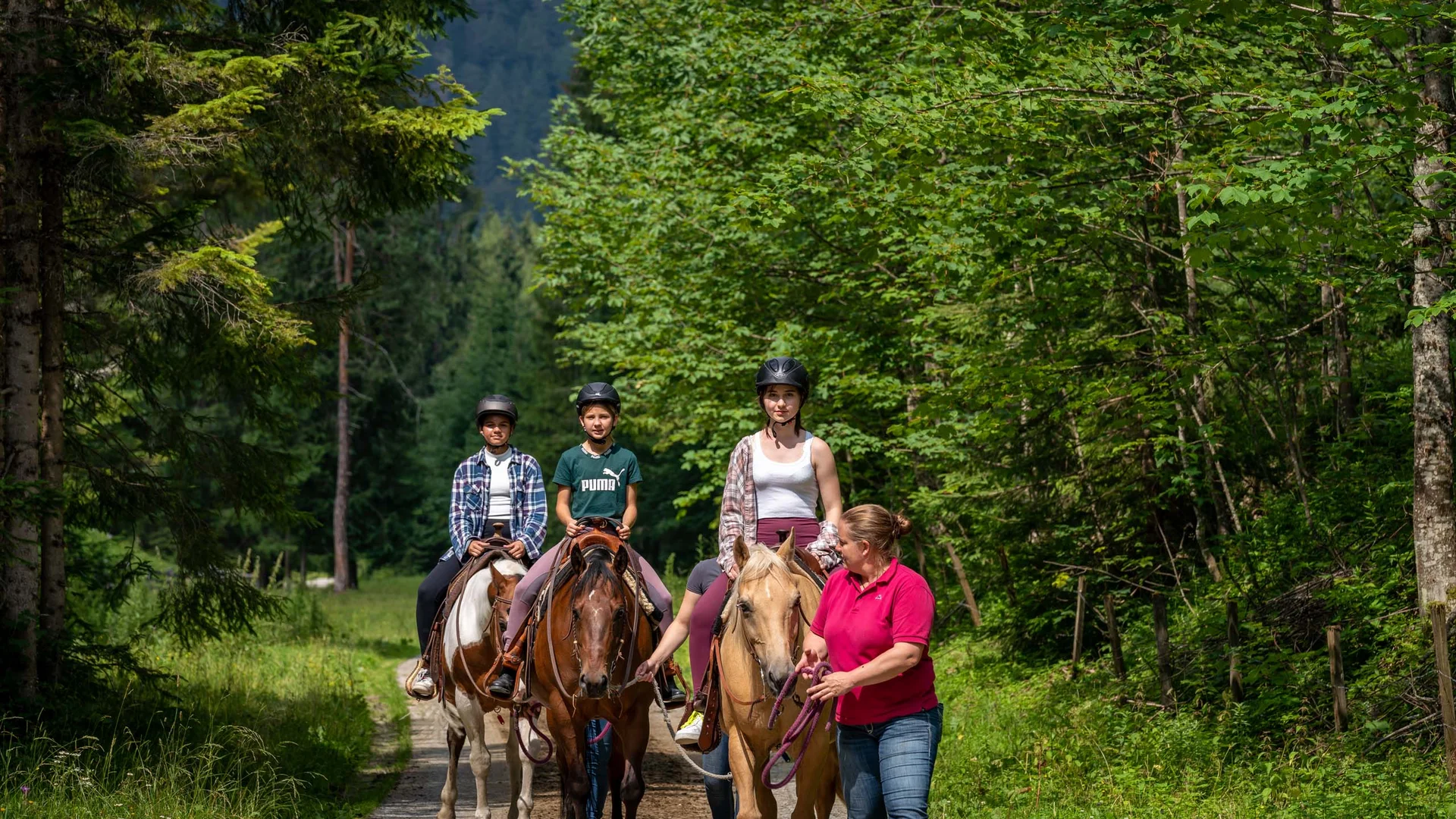 Familienhotel mit Ponyreiten am Achensee | Wiesenhof Pertisau Drei Reiter mit Helm auf Waldweg, geführt von einer Frau im roten Polohemd