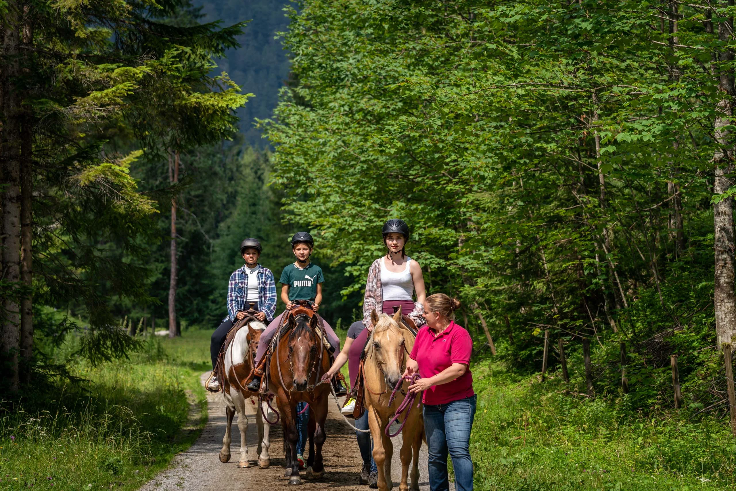 Familienhotel mit Ponyreiten am Achensee | Wiesenhof Pertisau Drei Reiter mit Helm auf Waldweg, geführt von einer Frau im roten Polohemd