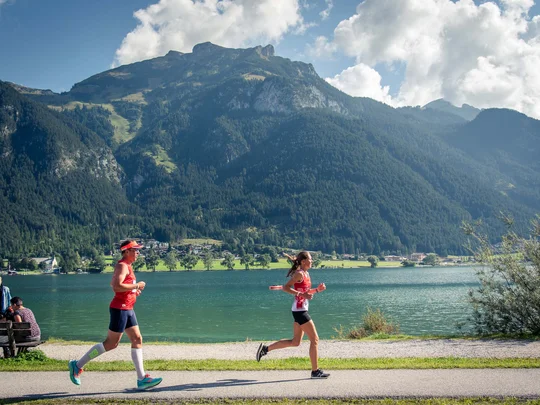 Zwei Personen joggen am See mit Bergen und Wolken im Hintergrund