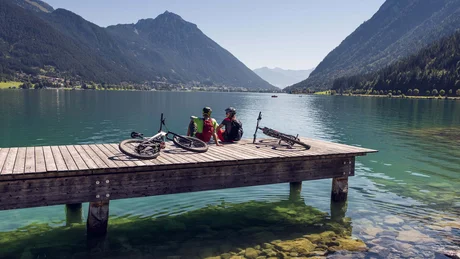 Zwei Radfahrer sitzen auf einem Steg am See mit Bergkulisse im Sonnenschein