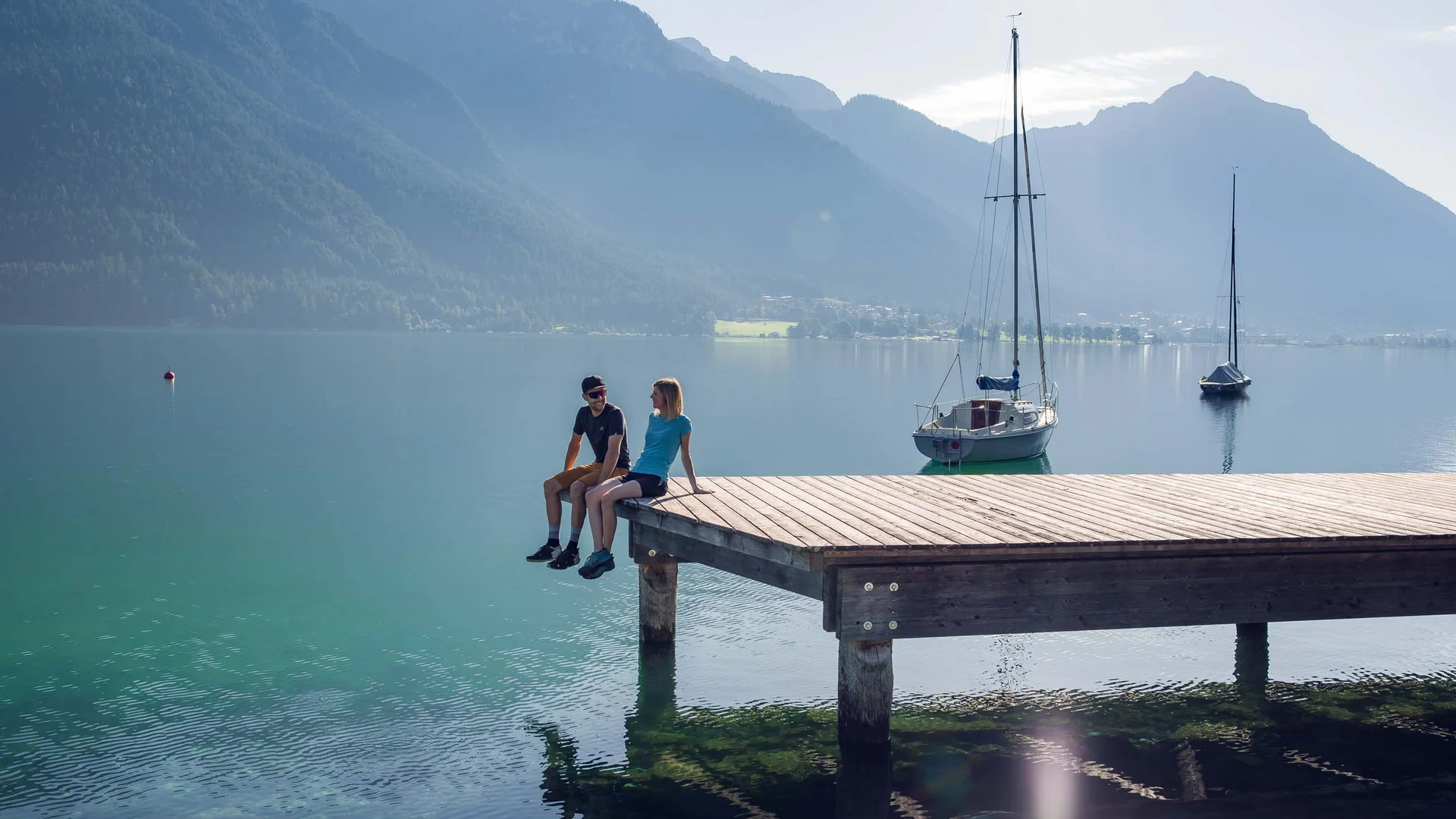In, auf und rund um den Achensee Zwei Menschen sitzen auf einem Steg über einem Bergsee mit Segelbooten im Hintergrund