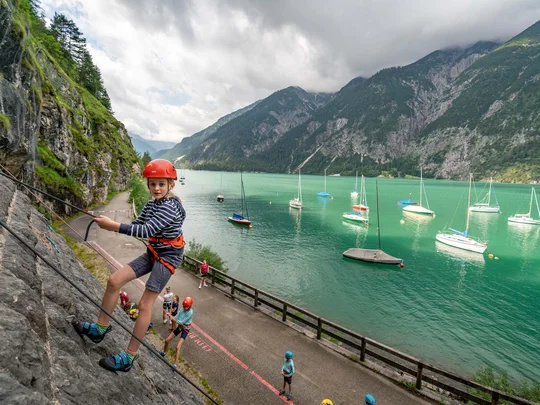 Sehenswürdigkeiten am Achensee rund um den Wiesenhof Kind beim Klettern an felsiger Steilwand mit Bergsee und Booten im Hintergrund