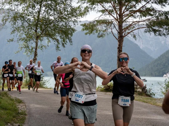 Läufer bilden Herz mit den Händen bei einem Rennen an einem See mit Bergen im Hintergrund