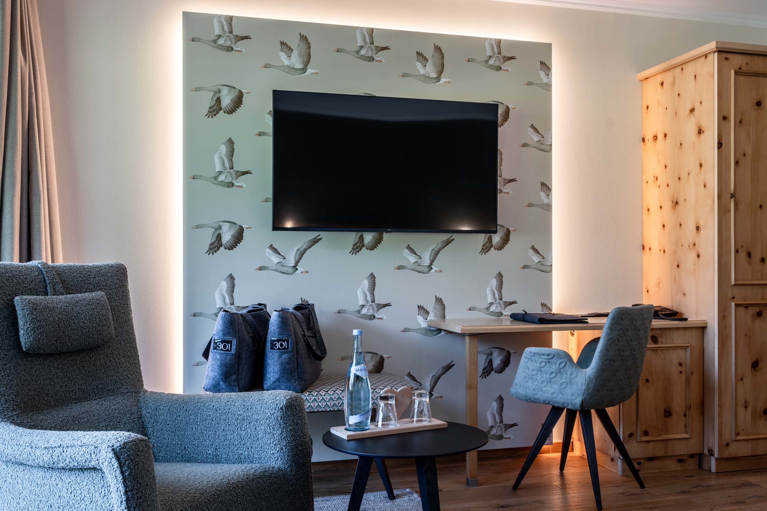Euer Familienhotel am Achensee Hotelzimmer mit TV, grauem Sessel, Schreibtisch und Vogel-Tapete
